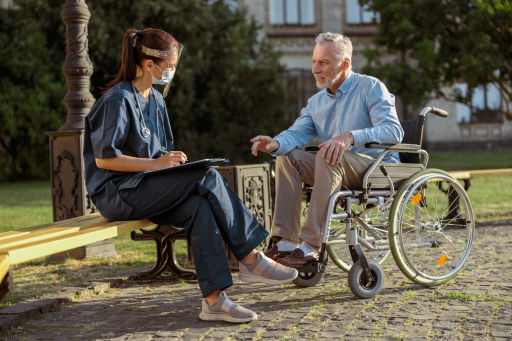 Full length shot of recovering male patient in wheelchair discussing his health condition with nurse