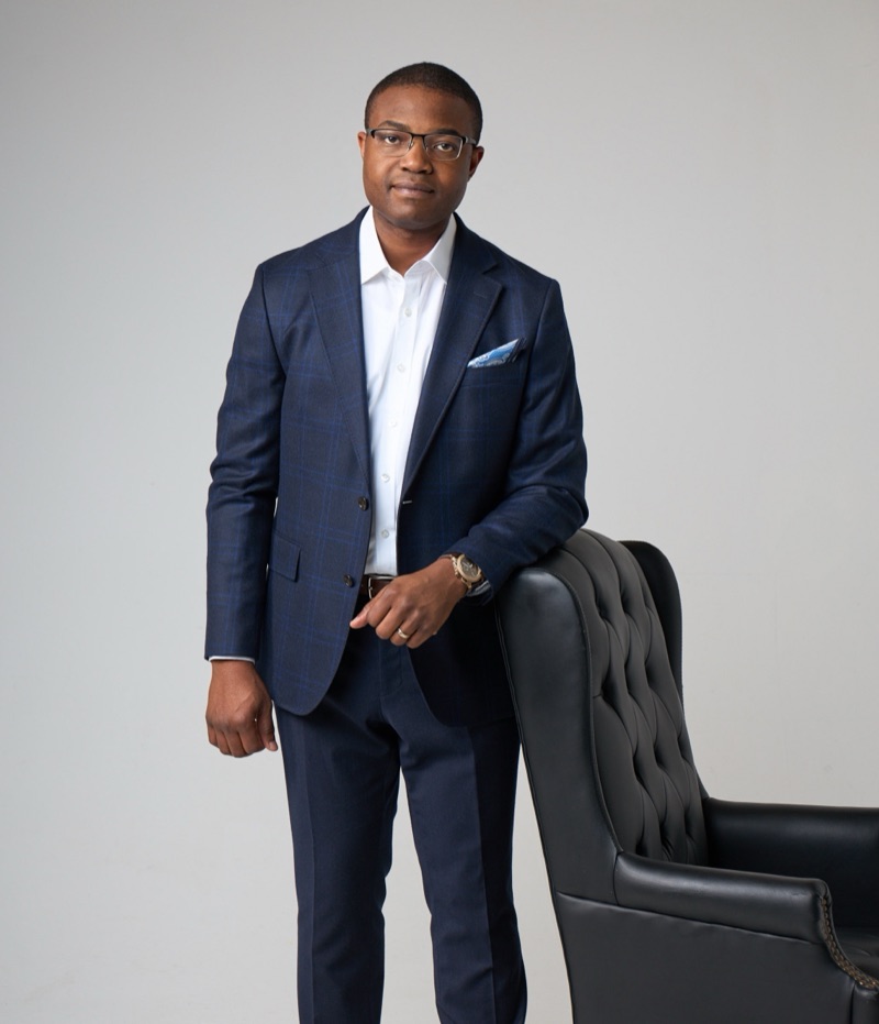 Abraham Oni standing beside a leather chair in a studio, hands clasped.