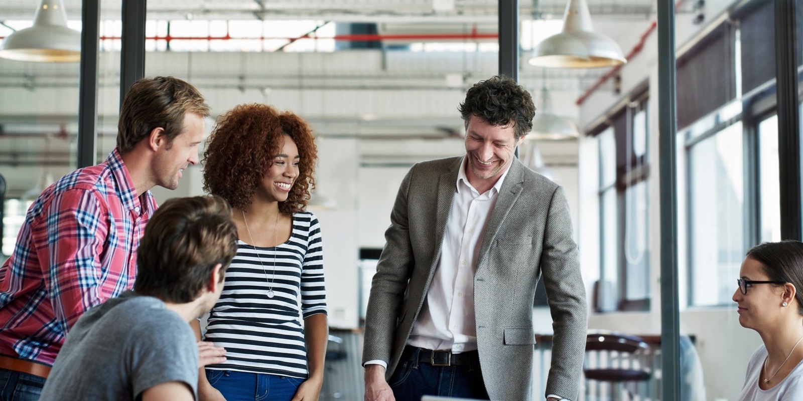 A cross-functional team standing around a table in a loft office, reviewing work together.